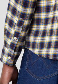 Geruite overhemd in geel, blauw en rood op een witte achtergrond, met een manchette met twee knopen en een gebogen zoom, gecombineerd met denim.