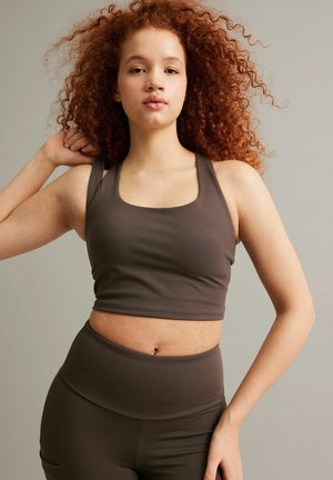 Junge Frau mit lockigem, rotem Haar, die einen dunkelbraunen Sport-BH und hoch taillierte Leggings trägt, posiert vor einem grauen Hintergrund.