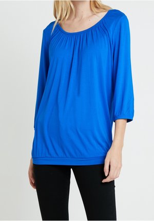 Femme portant un chemisier ample bleu vif à manches trois-quarts et un pantalon noir, debout devant un fond clair uni.
