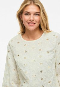 Femme blonde souriante, portant un chemisier blanc en dentelle œillets avec des motifs floraux et de petites boucles d'oreilles en or contre un fond uni.
