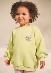 Light green sweatshirt with puffed sleeves, featuring a decorative patch of a smiling apple in pastel colors. Soft fabric and a relaxed fit.