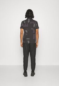 Polo à manches courtes noir avec motifs abstraits gris. Associé à un pantalon noir fuselé et des chaussures épaisses noires. Fond uni.