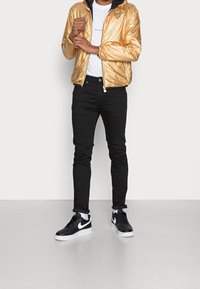 Zlatá kovová bunda na zip s černou kapucí, bílé tričko, černé džíny a černé tenisky Nike. Na bundě je barevné logo.