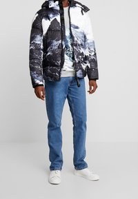 Prošívaná bunda s potiskem hor v černé, bílé a modré barvě, se zipem, nastavitelnou kapucí, doplněná šedou mikinou s potiskem a modrými džínami.