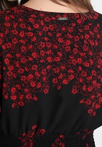 Schwarzer Stoff mit einem roten Blumenmuster, das kleine Rosen und grüne Stängel zeigt. Elastischer Bund mit einem Etikett am Halsausschnitt.