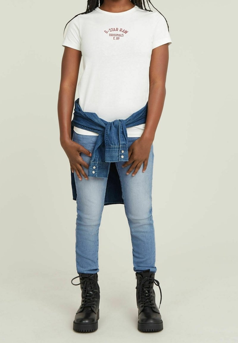 Barn som bär en vit T-shirt med grafik, ljusblå jeans, svarta snörkängor, och en jeansjacka knuten runt midjan, står mot en enfärgad bakgrund.