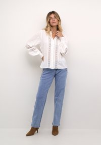 Vit skjorta med knappar och spetsdetaljer längs framsidan och ärmarna, kombinerad med ljusblå raka jeans och bruna klackade ankelskor.