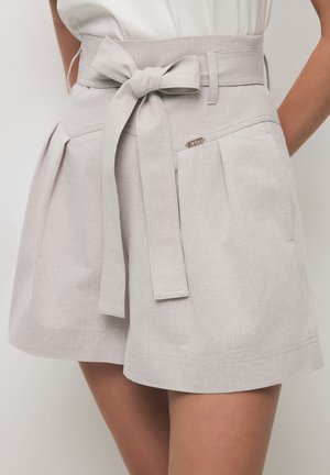 Beige short met hoge taille, plooien en een stoffen riem die is gestrikt, gedragen met een wit topje dat is ingestopt, waardoor een deel van de taille en dijen zichtbaar is.