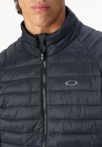 Wypełniona czarna kurtka z wysokim kołnierzem, pikowanym wzorem i zamkiem z przodu. Posiada logo po lewej stronie klatki piersiowej.