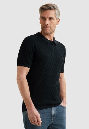 Mann mit grauen Haaren, der ein schwarzes strukturiertes kurzärmeliges Poloshirt und blaue Jeans trägt, steht mit einer Hand in der Tasche und blickt zur Seite.