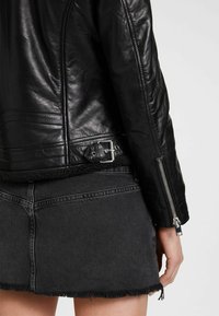 Zwarte leren jacket met een textuurafwerking, zijrits en gespsluiting bij de taille, gecombineerd met een versleten zwarte denim rok.