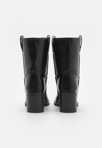 Un par de botas de cuero negro hasta la mitad de la pantorrilla con tacones bloque apilados, vistas desde atrás sobre un fondo blanco.