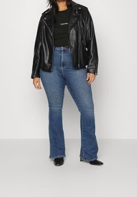 Svart fusk läder bikerjacka över en mörkgrön t-shirt, tillsammans med högmidjade blå utsvängda jeans och svarta klackstövlar.