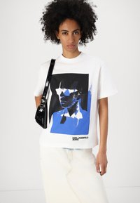 Biela bavlnená tričká s odvážnou grafickou potlačou tváre v čiernej a modrej farbe. Obsahuje text "KARL LAGERFELD JEANS" na spodnej časti.