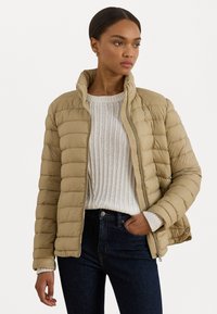 Giacca puffa beige con collo alto, design a trapunta e chiusura con zip. Indossata sopra un maglione bianco lavorato con tessuto testurizzato e jeans scuri.
