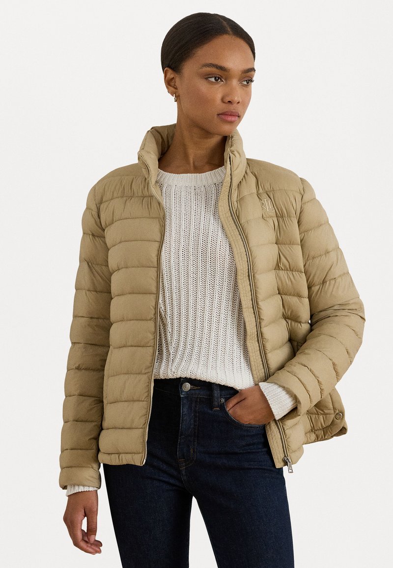 Giacca puffa beige con collo alto, design a trapunta e chiusura con zip. Indossata sopra un maglione bianco lavorato con tessuto testurizzato e jeans scuri.