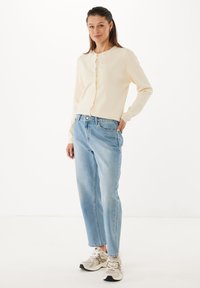 Kvinna som står i krämfärgad kofta, ljusblå jeans och beige sneakers mot vit bakgrund, ler med handen på höften.