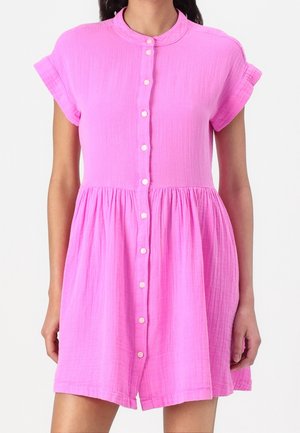Różowa sukienka na guziki z krótkim rękawem, z marszczonym taliowaniem i stójką, noszona przez stojącą osobę na jednolitym tle.