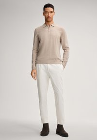 Beige Langarm-Polo-Pullover mit einem Reißverschluss am Kragen, kombiniert mit schmal geschnittenen weißen Hosen und dunkelbraunen Stiefeletten. Glatte Textur.