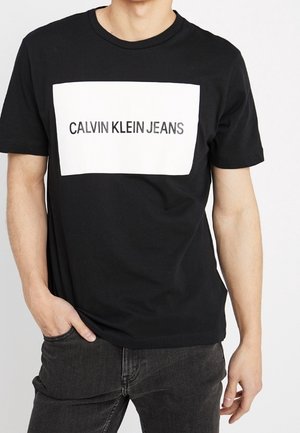 Mand iført en sort T-shirt med hvidt rektangulært logo med teksten "CALVIN KLEIN JEANS" og mørke jeans, hånden i lommen, foran ensfarvet baggrund.