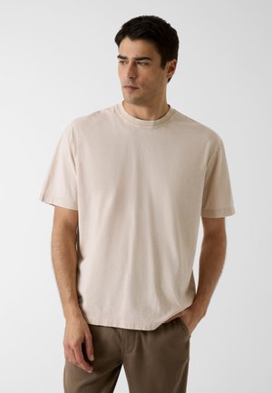 Man met kort donker haar, gekleed in een beige T-shirt met korte mouwen en een bruine broek, kijkt naar links tegen een effen achtergrond.