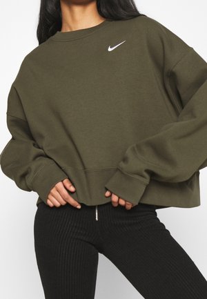 Kobieta w oversize oliwkowej zielonej bluzie Nike z długimi rękawami i czarnych prążkowanych legginsach z przednim zamkiem.