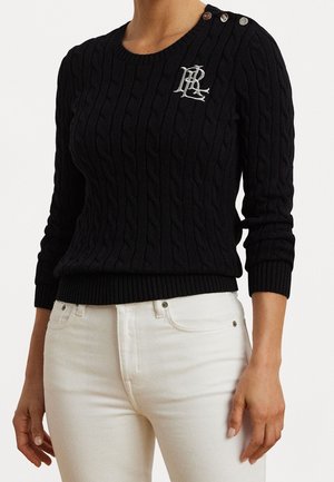 Pull noir en maille torsadée avec logo brodé blanc sur la poitrine et détails à boutons sur l'épaule, porté avec un pantalon blanc.