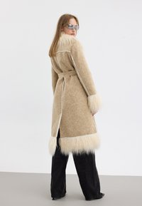 Manteau beige avec une finition texturée, col et poignets en fausse fourrure, ouvert à l'avant, taille ceinturée et fentes sur les côtés, associé à un pantalon noir.