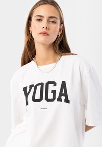 Camiseta blanca de algodón, con un atrevido estampado en negro de "YOGA" y un pequeño logo. Corte relajado, mangas cortas y escote redondo.