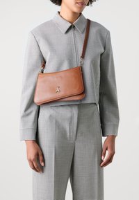 Borsa a tracolla in pelle marrone con una texture liscia, forma rettangolare, emblema d'ape argentata e tracolla regolabile, indossata sopra una camicia a quadri.