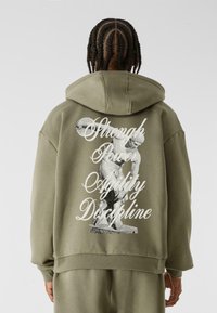 Sweat à capuche vert olive avec un design graphique d'une statue et un texte blanc indiquant "Force, Puissance, Agilité, Discipline" au dos.