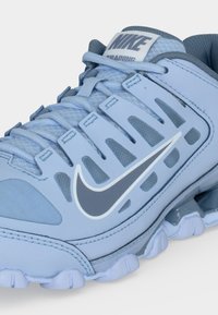 Ανοιχτό μπλε αθλητικό παπούτσι προπόνησης Nike με γκρι λογότυπο swoosh, επάνω μέρος από δίχτυ και ανθεκτική σόλα, απεικονίζεται σε κοντινή γωνιακή όψη.