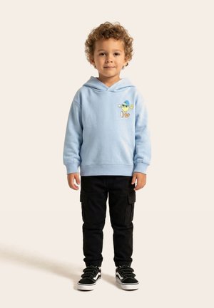 Băiat tânăr cu păr creț purtând hanorac albastru deschis cu patch desen animat, pantaloni cargo negri și adidași negri, stând în picioare pe un fundal bej.