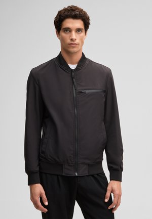 Junger Mann mit kurzen lockigen Haaren, der eine schwarze Bomberjacke mit Reißverschluss und einer Reißverschlusstasche auf der Brust trägt, steht vor einem schlichten hellen Hintergrund.