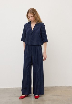 Femme portant un costume bleu marine à rayures fines avec un pantalon à jambes larges et un blazer à manches courtes, debout et regardant vers le bas, portant des chaussures plates rouges.