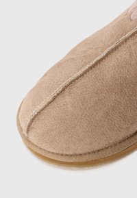 Närbild av en beige ulltoffel med synliga sömmar och en gummisula på en vit bakgrund.