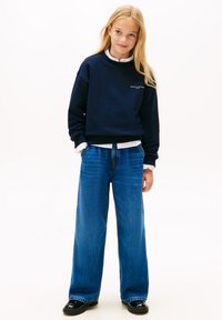 Felpa blu con polsini e orlo a coste, abbinata a jeans a gamba larga blu. L'outfit presenta una camicia bianca con colletto sotto.