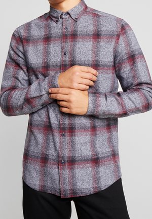 Mand iført en blød flannelskjorte med lange ærmer i gråt, rødt og sort ternet mønster, knappet op, justerer venstre manchet.