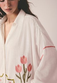 Nainen, joka pukeutunut valkoiseen teksturoituun puseroon, jossa on etuosassa vaaleanpunaisia kirjottuja tulppaaneja ja hihoissa punaiset raidat, taustalla yksivärinen beige.