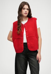 Sarkans vilnas bezdelīgs ar teksturētu apdari, aprīkots ar apaļu kakla izgriezumu, pogām ar spiedpogām un priekšējām kabatām. Nēsāts virs baltas T-kreklu.