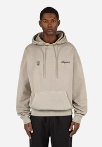 Gaisas bēšas kokvilnas kapucēta džemperis ar priekšējo kabatu, sasaistes kapuci un izšūtu logo uz krūšu laukuma. Brīva piegriezuma dizains.