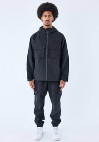 Giacca nera impermeabile con due grandi tasche frontali e chiusura zip, abbinata a pantaloni cargo coordinati e sneakers bianche.