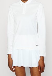 Vit långärmad polo-topp med krage och knäppning. Nedan en matchande plisserad kjol. Litet svart Nike-logga på topens sida.