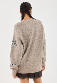 Pull beige surdimensionné en tissu maille doux avec des motifs de chats noirs sur les manches et un ourlet côtelé. Porté sur une jupe sombre à couches.