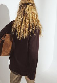 Mørkebrun, overdimensioneret skjorte med lange ærmer og knaplukninger; person holder en beige ruskinds taske. Lys baggrund og bløde skygger.