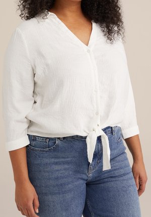 Vrouw met een gestructureerde witte blouse met knopen, vastgeknoopt in de taille, driekwart mouwen en blauwe spijkerbroek.