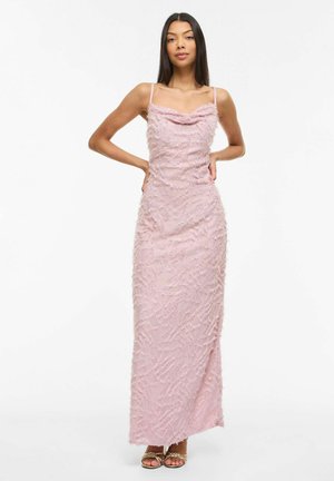 Donna in piedi con le mani sui fianchi che indossa un abito lungo rosa senza maniche e con tessuto a trama, e sandali dorati con punta aperta, su sfondo bianco.