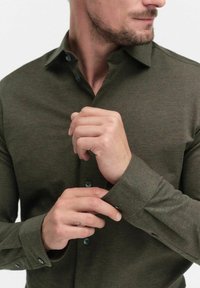 Chemise à manches longues vert olive avec col boutonné, tissu texturé et boutons bleus contrastants aux poignets et à la patte de boutonnage.