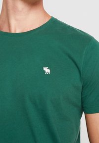 Grünes T-Shirt mit kurzen Ärmeln aus weichem Baumwollstoff, mit einem kleinen weißen Elch-Logo auf der Brust. Klassisches Crewneck-Design.