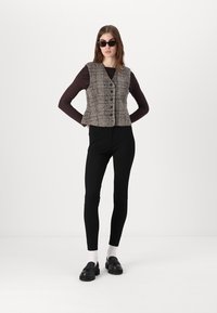 Brūns un melns raibs veste ar priekšējiem pogām, uzvilkta virs garām piedurknēm, apvienota ar melnām legingām un melnām kurpēm. Minimālisma dizains.